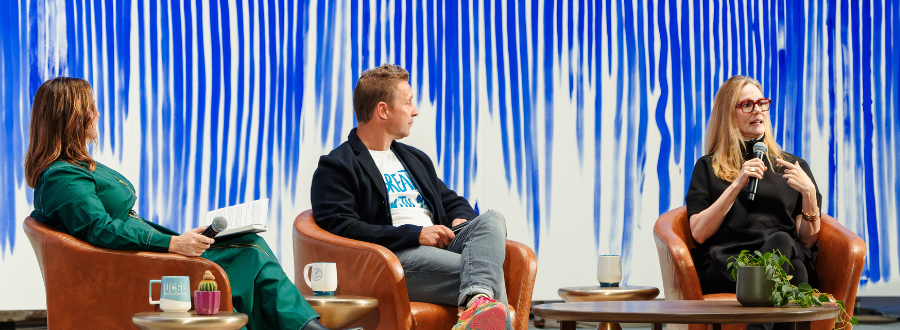 8 of 8, Three adults sit in chairs on a stage, behind them are white canvases covered in blue lines. they're having a conversation and holding microphones, all are white, two are women and one is a man. 